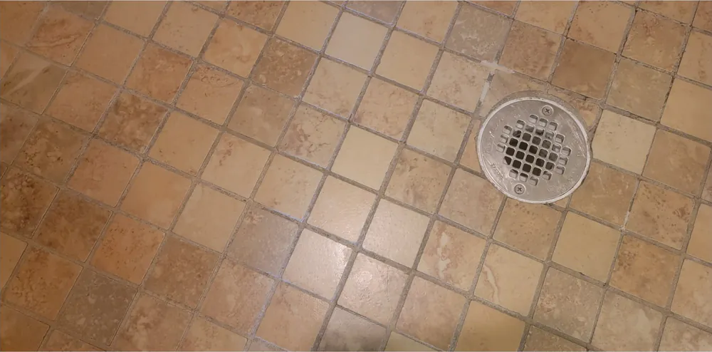 Before: a small tan tile shower floor with dark grout around the drain.
